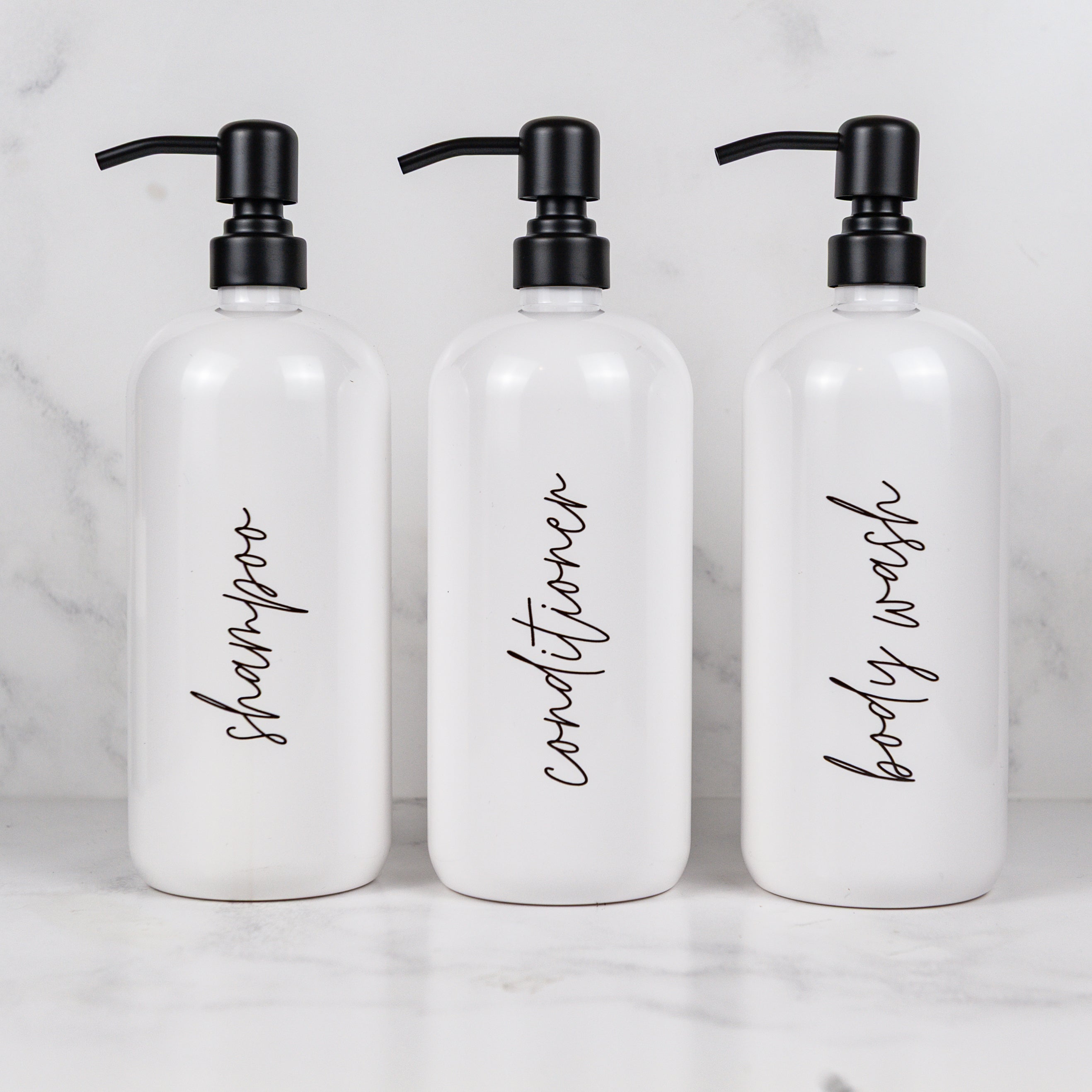 Three white bottles labeled 'shampoo', 'conditioner', and 'body wash' on a marble surface.