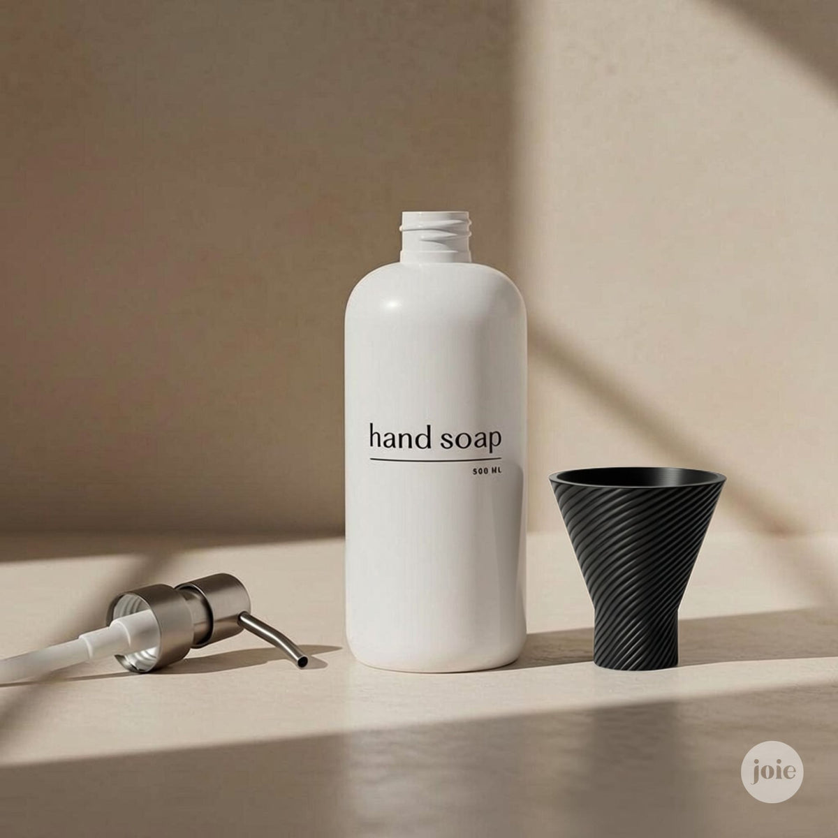 A matte white hand soap bottle labeled 'hand soap' stands on a beige surface, with a black funnel-shaped container and a silver-and-white pump dispenser placed beside it in warm, directional light.