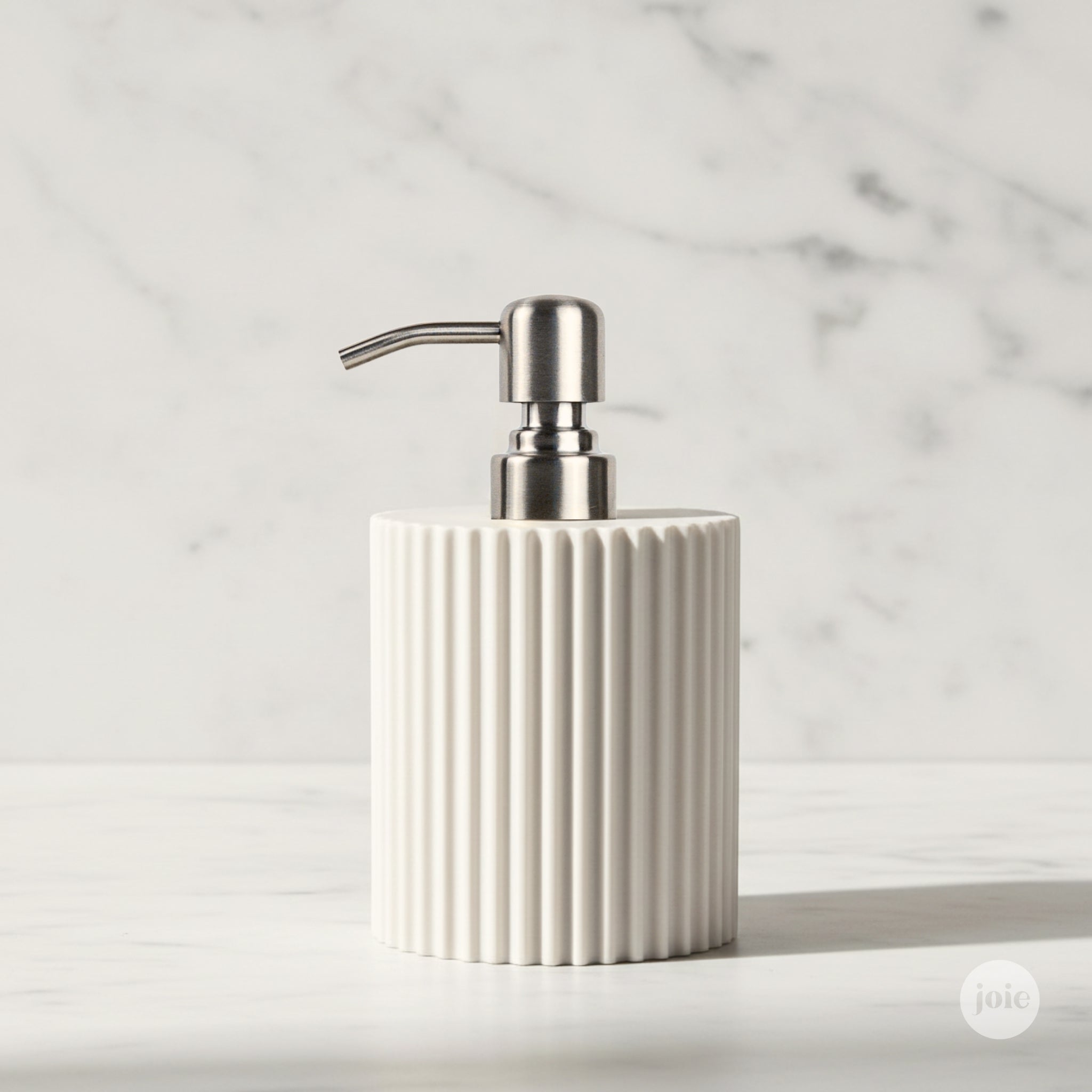 A ribbed ceramic soap dispenser in warm off-white with a brushed silver metal pump, set against a white marble background.