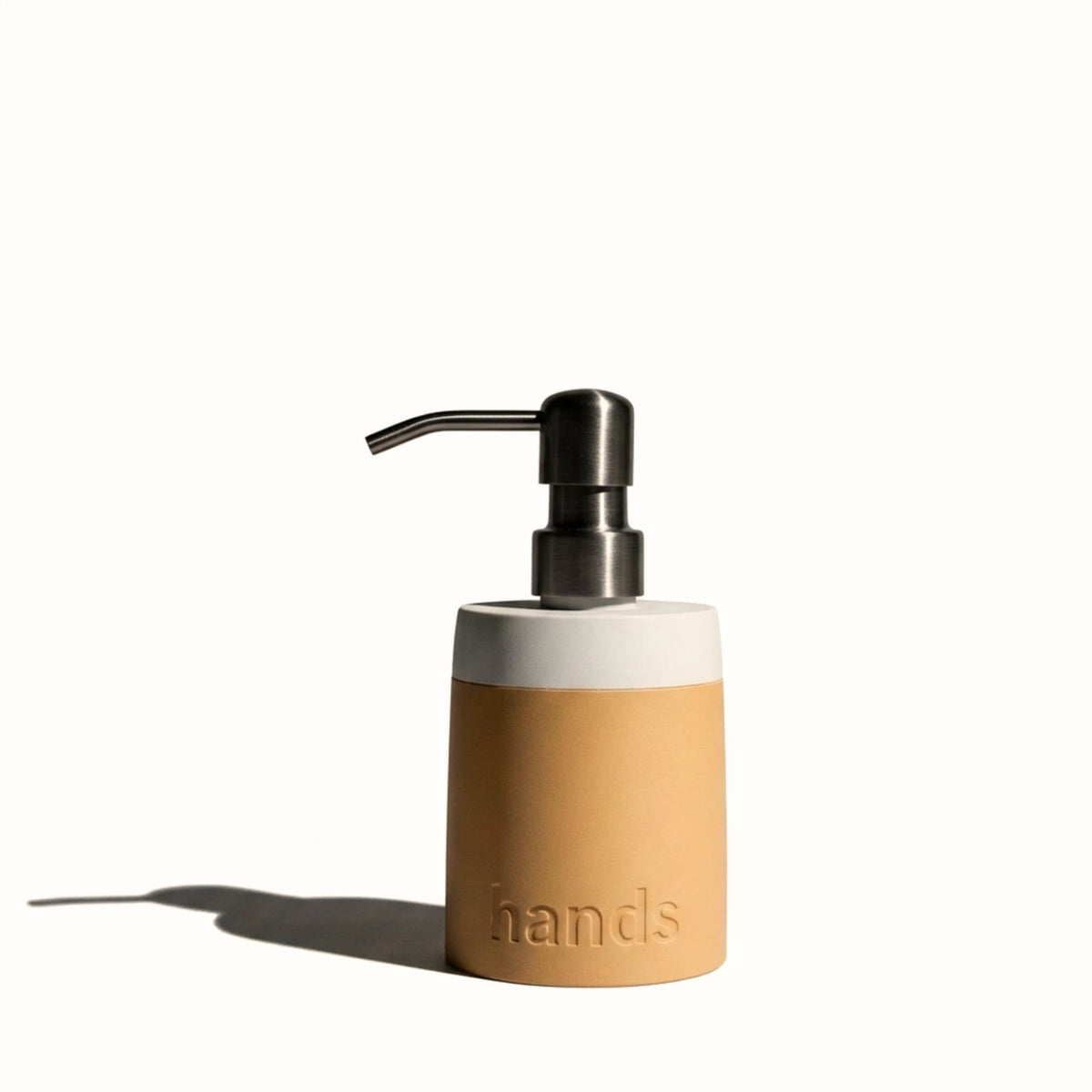 A cylindrical hand sanitizer dispenser with a warm tan textured lower body, a white upper band, and a dark brushed metal pump, shown on a white background with a soft shadow.