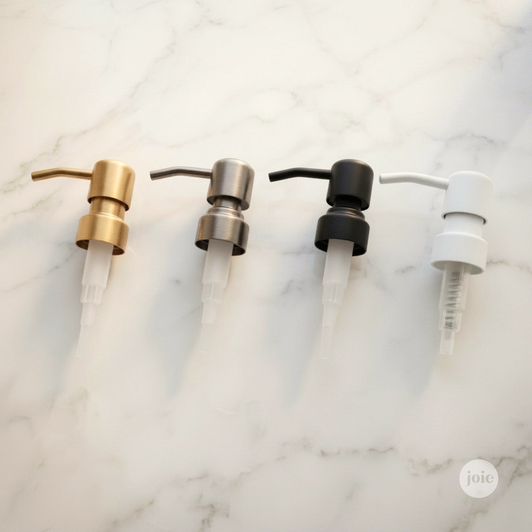 Four pump dispenser heads in gold, silver, black, and white finishes arranged in a row on a white marble background.