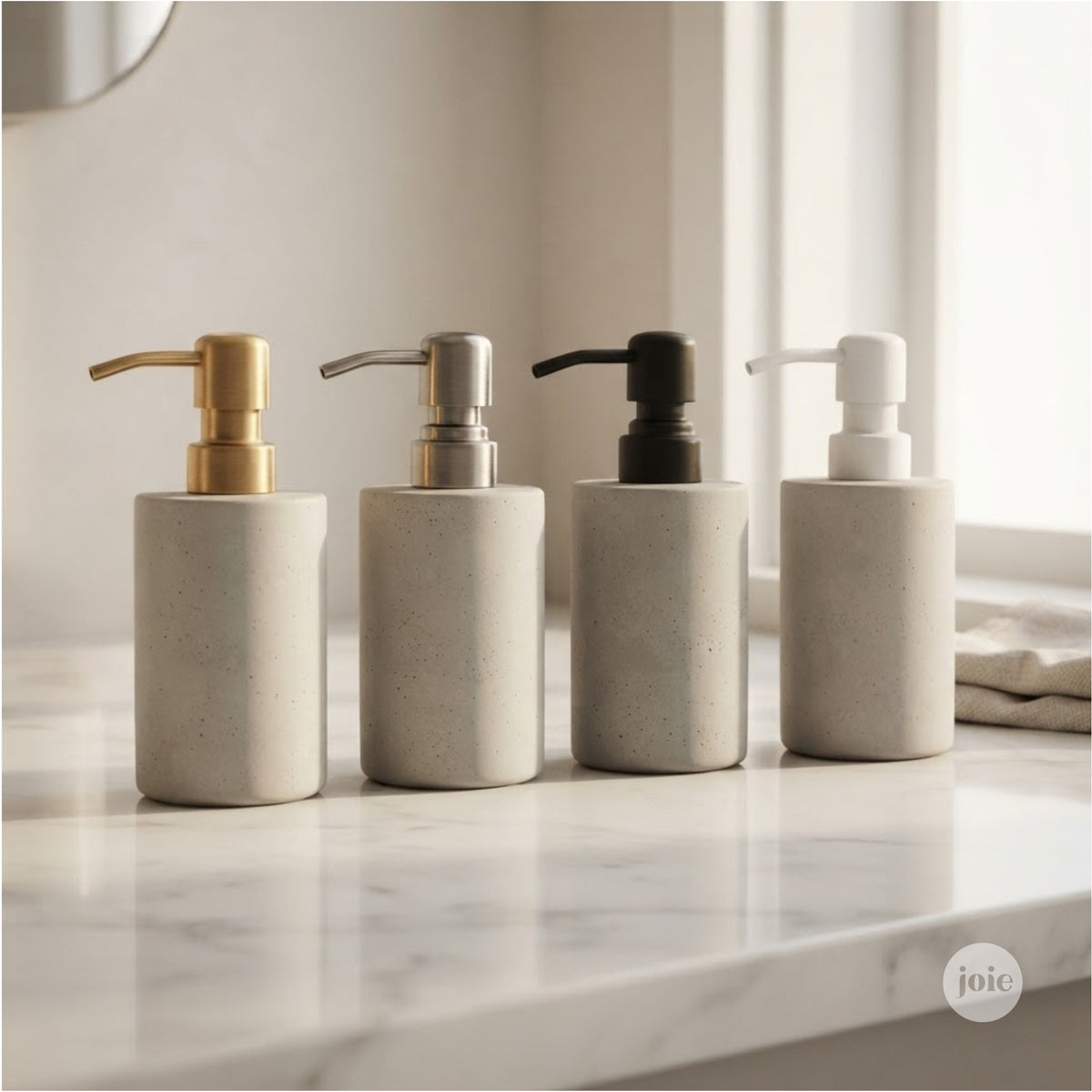 Four cylindrical soap or lotion dispensers with textured light stone bases and pump tops in gold, silver, black, and white metal, lined up on a white marble countertop.