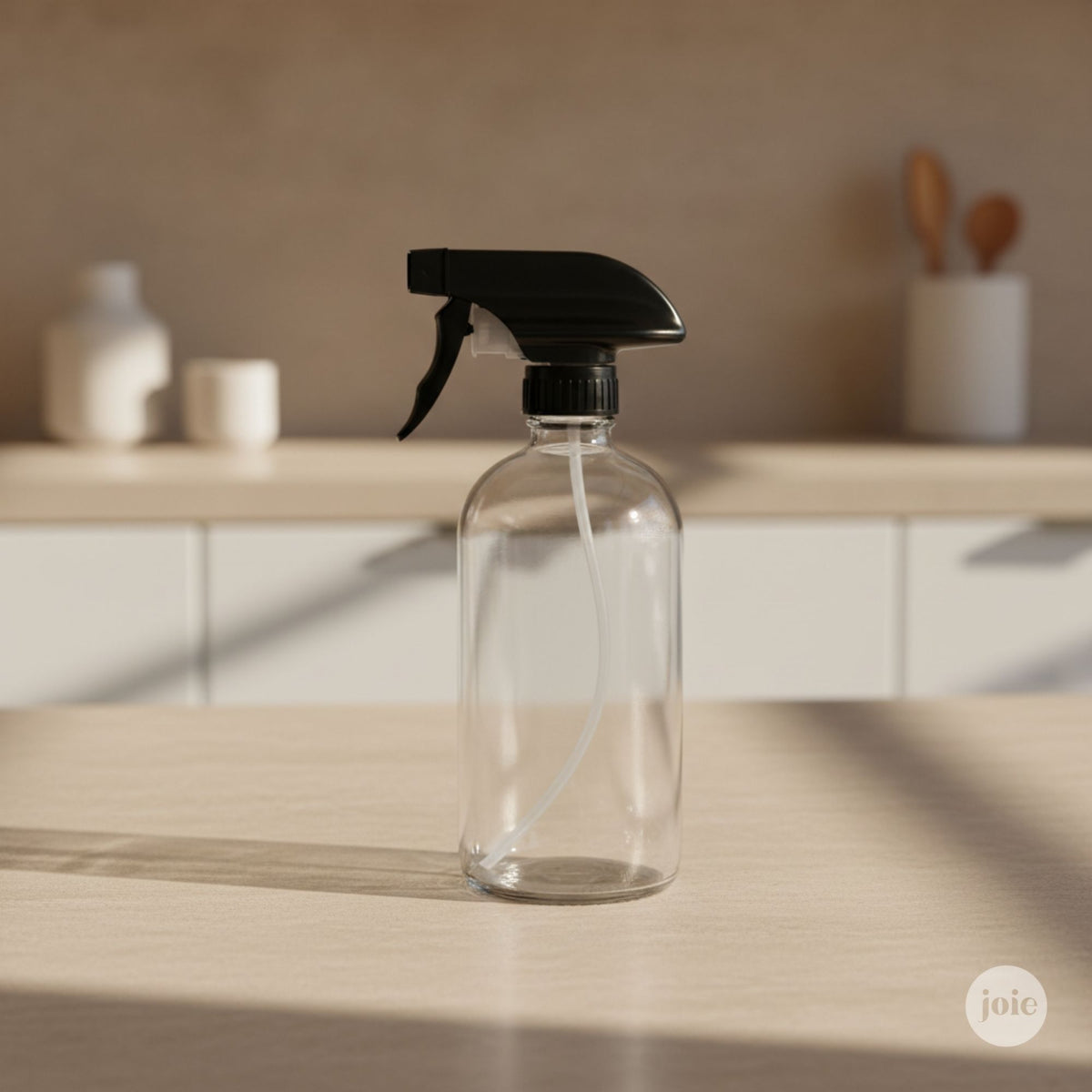 A clear glass spray bottle with a black trigger sprayer, standing on a light wood countertop in a softly blurred kitchen setting.