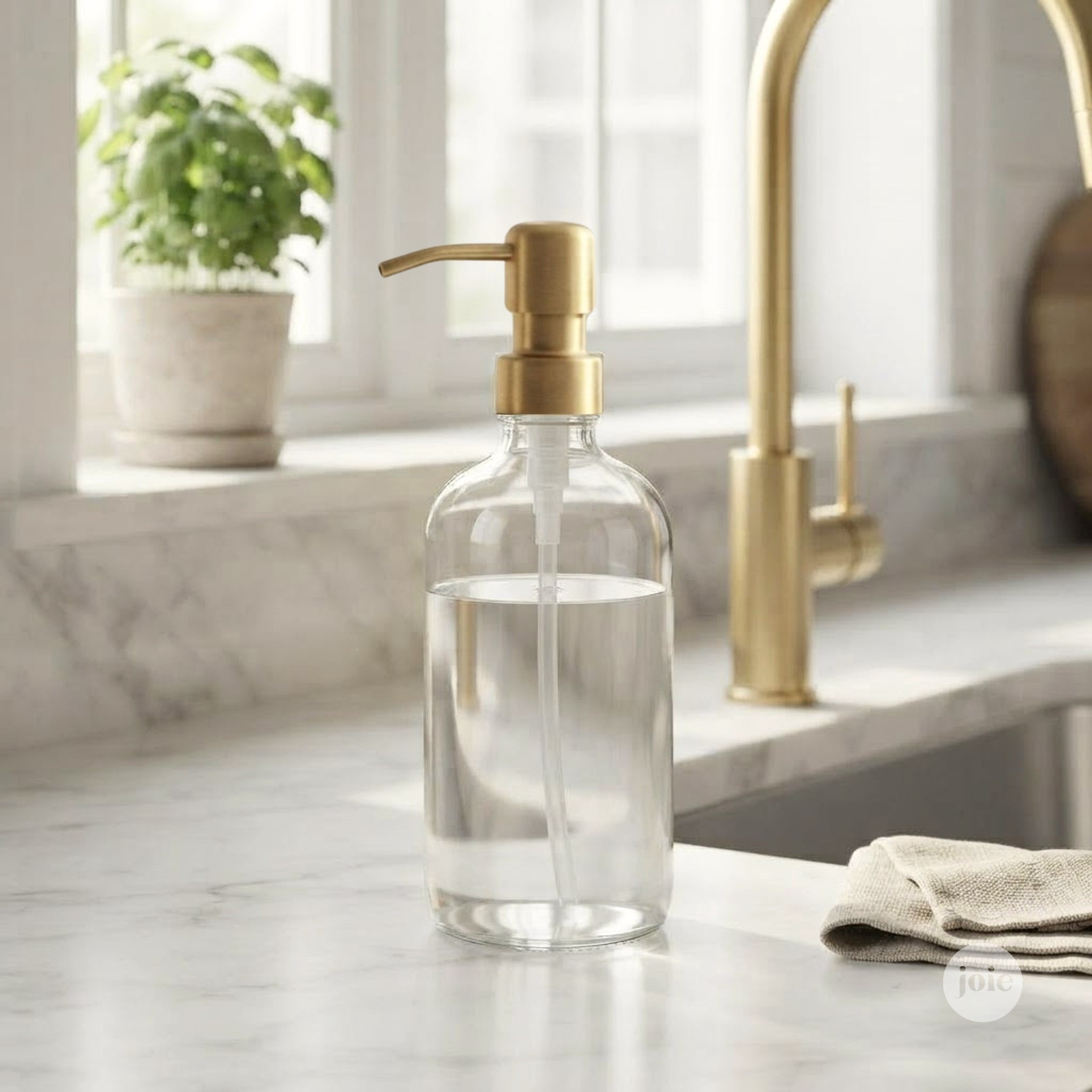 A clear glass pump bottle with a brushed gold dispenser, filled with soap, set on a white marble kitchen countertop near a gold faucet.