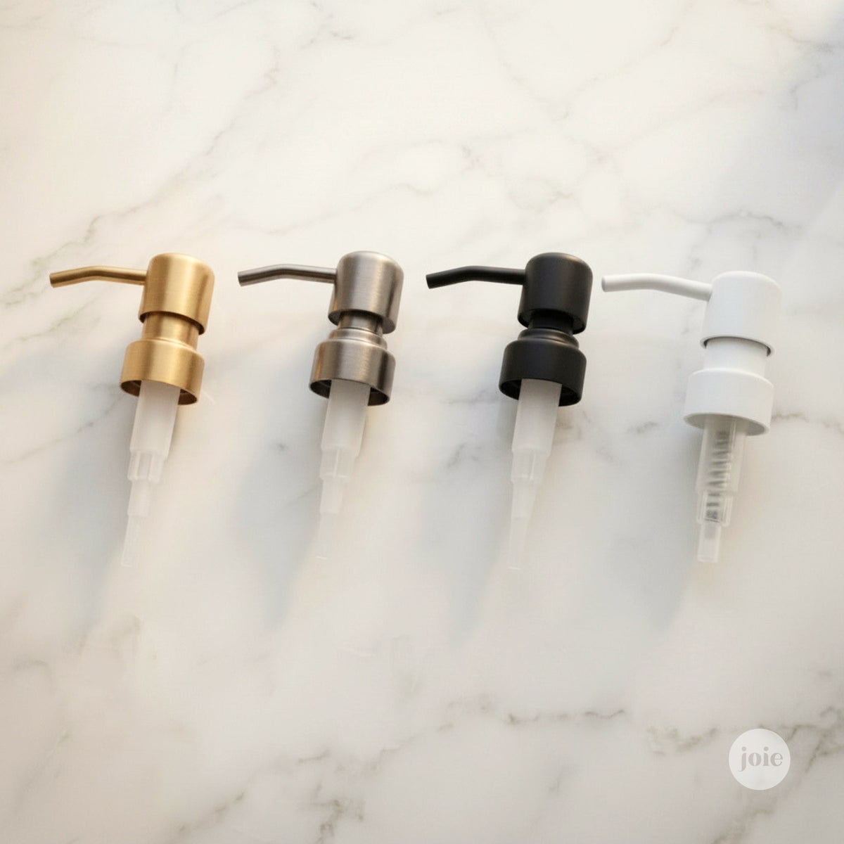 Four pump dispenser heads in gold, silver, black, and white finishes arranged in a row on a white marble background.