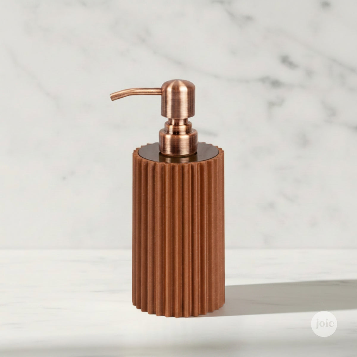 A modern cylindrical soap dispenser with a ribbed copper-toned body and a matching bronze metallic pump, set against a white marble background.