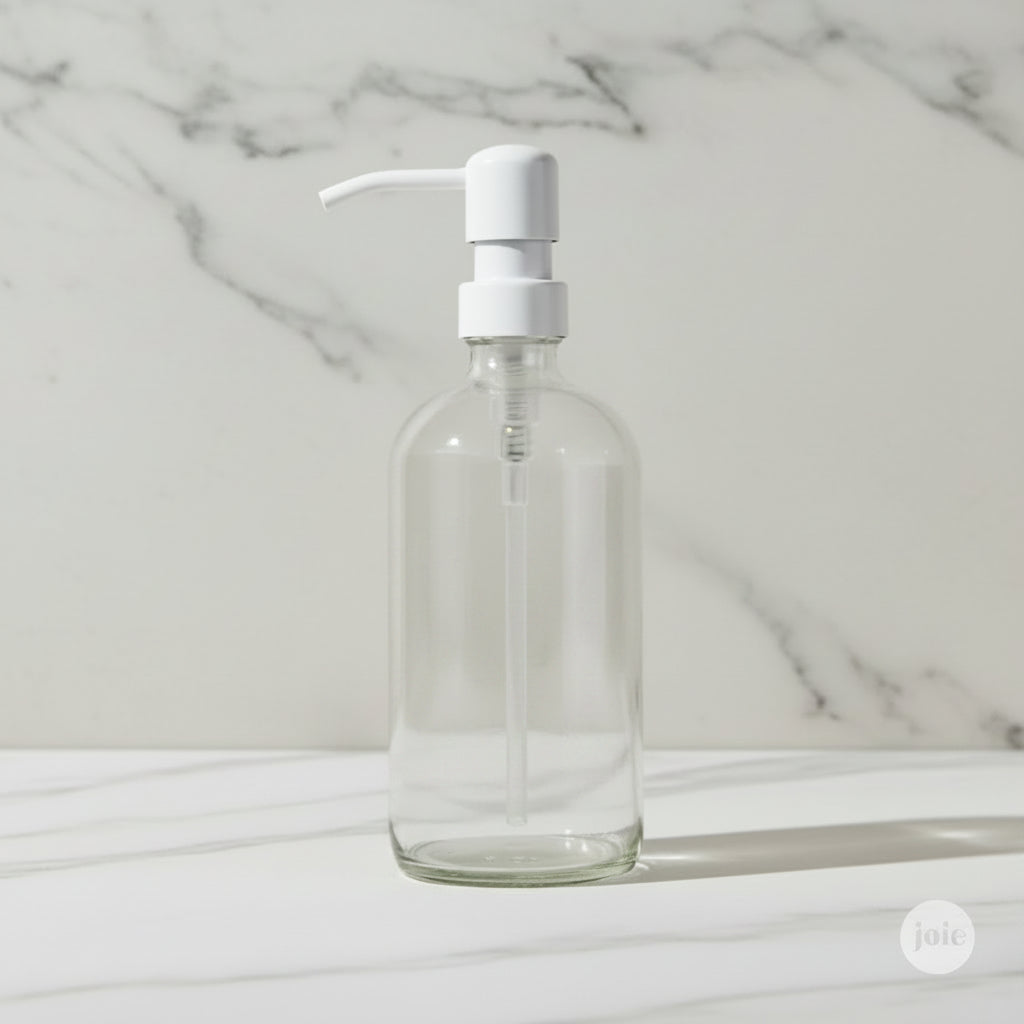 A clear glass hand soap dispenser bottle with a premium white metal pump set against a neutral light marble background.