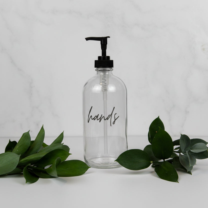 A clear glass soap dispenser bottle with the word 'hands' written on it.