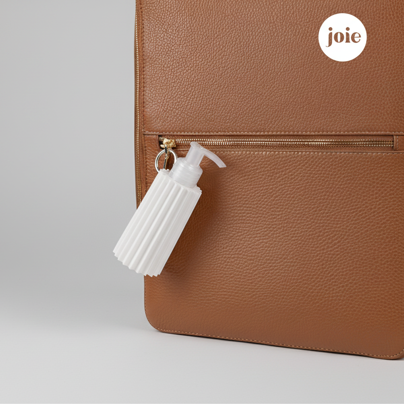 Brown leather bag with a white travel size lotion pump and 'joie' brand logo on a light gray background